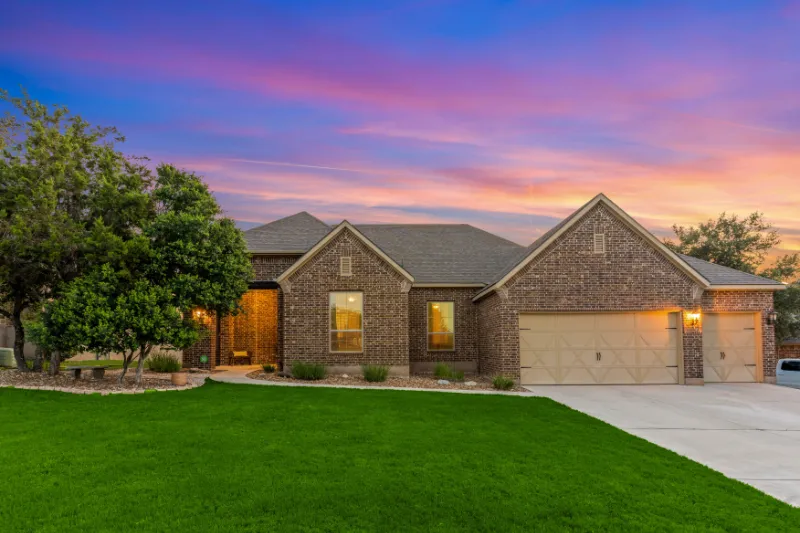 Single-story brick house with a three-car garage, lit windows, and a well-kept green lawn, set against a colorful sunset sky with pink and purple hues. Trees and shrubs line the front walkway.
