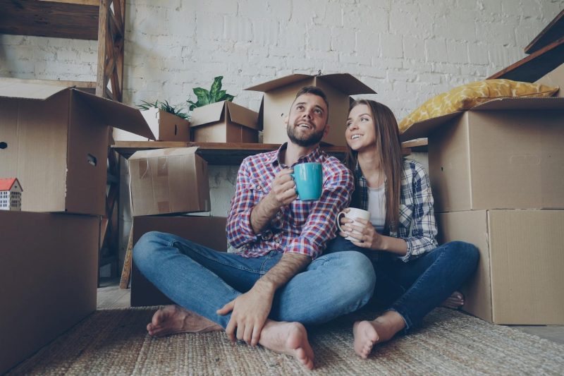 Couple sitting on the floor with coffee, surrounded by unpacked moving boxes in their new home.