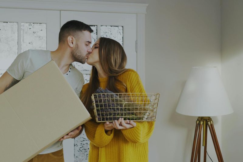 A couple kisses while holding a moving box and a basket in their new home.