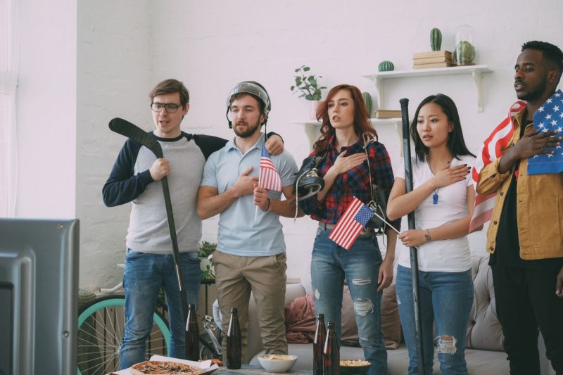Diverse friends gather indoors, watching sports on TV surrounded by American flags.