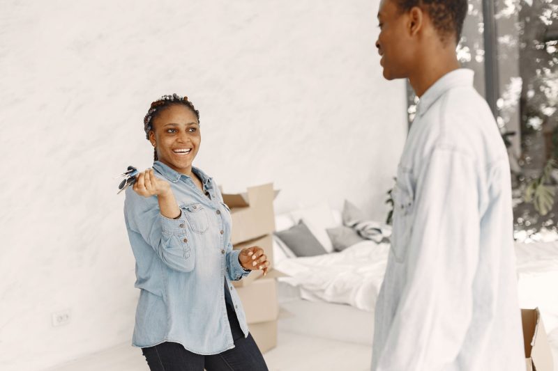 Smiling couple indoors, holding house keys, with a cozy living space in the background.